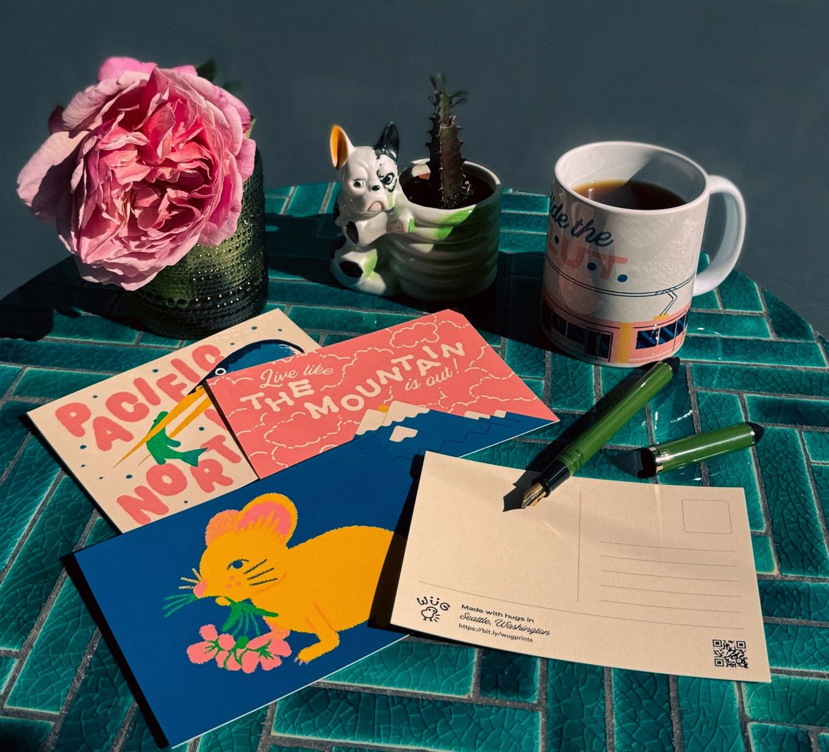 Wug Prints postcards arranged on a table with a mug of coffee and a fresh flower