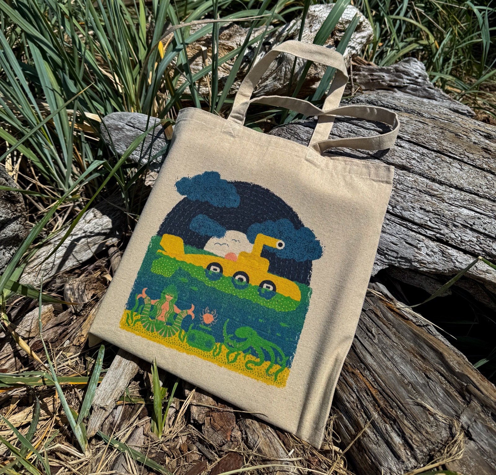 A Wug Prints tote bag featuring an illustrated design, resting on driftwood